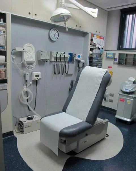 Fig 1. Single-chair aesthetic procedure room: reclining couch, orientable overhead cold light, wall-mounted diagnostic instruments and monitors, clock, and a benchtop steriliser at right, on a seamless sealed vinyl floor.